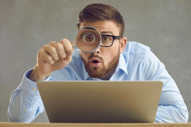 Mann sitzt vor einem Notebook und kontrolliert mit einer Lupe, was er am Bildschirm sieht
